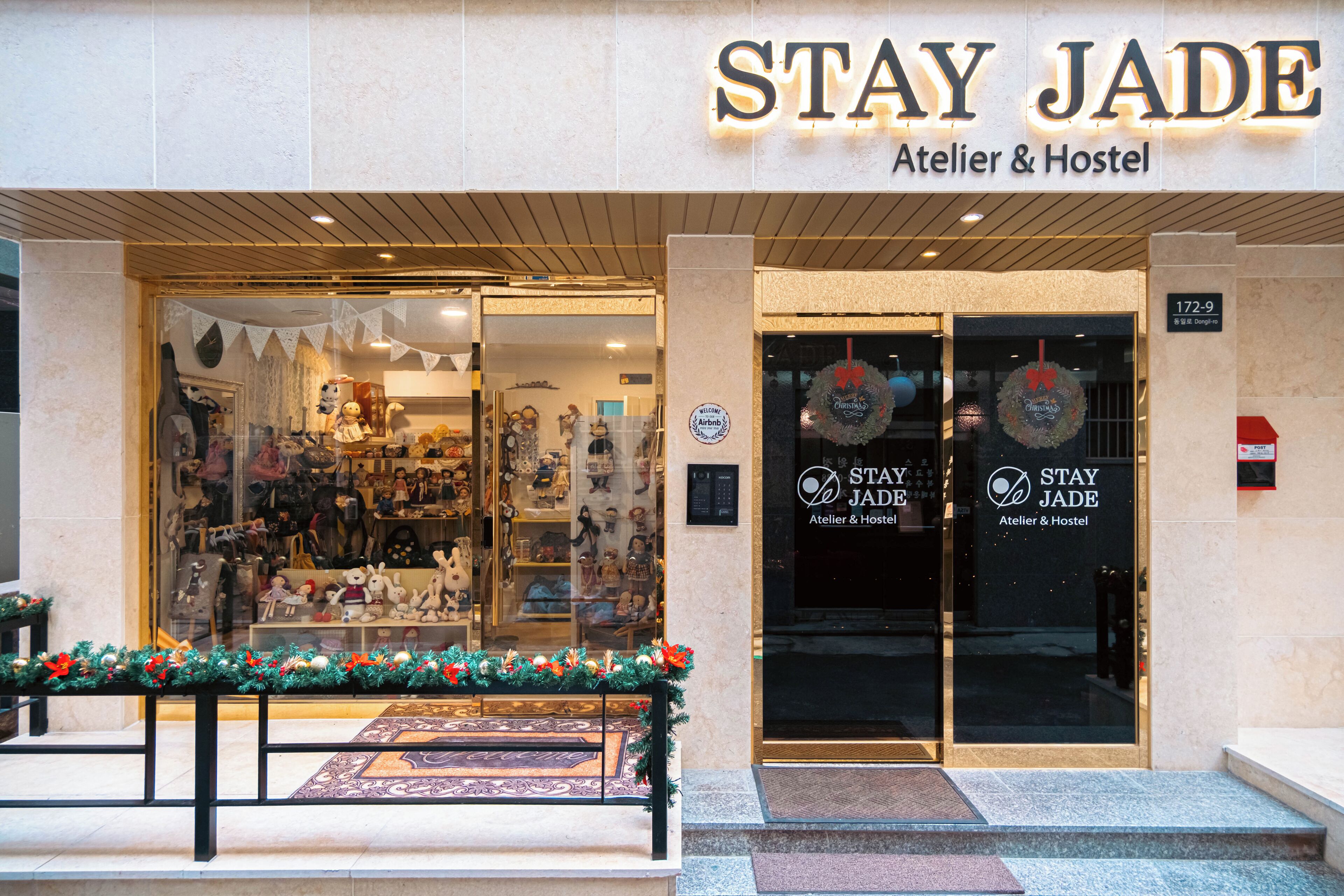 Exterior del Stay Jade, un Atelier & Hostel con entrada de vidrio, decoraciones navideñas y un banco afuera.