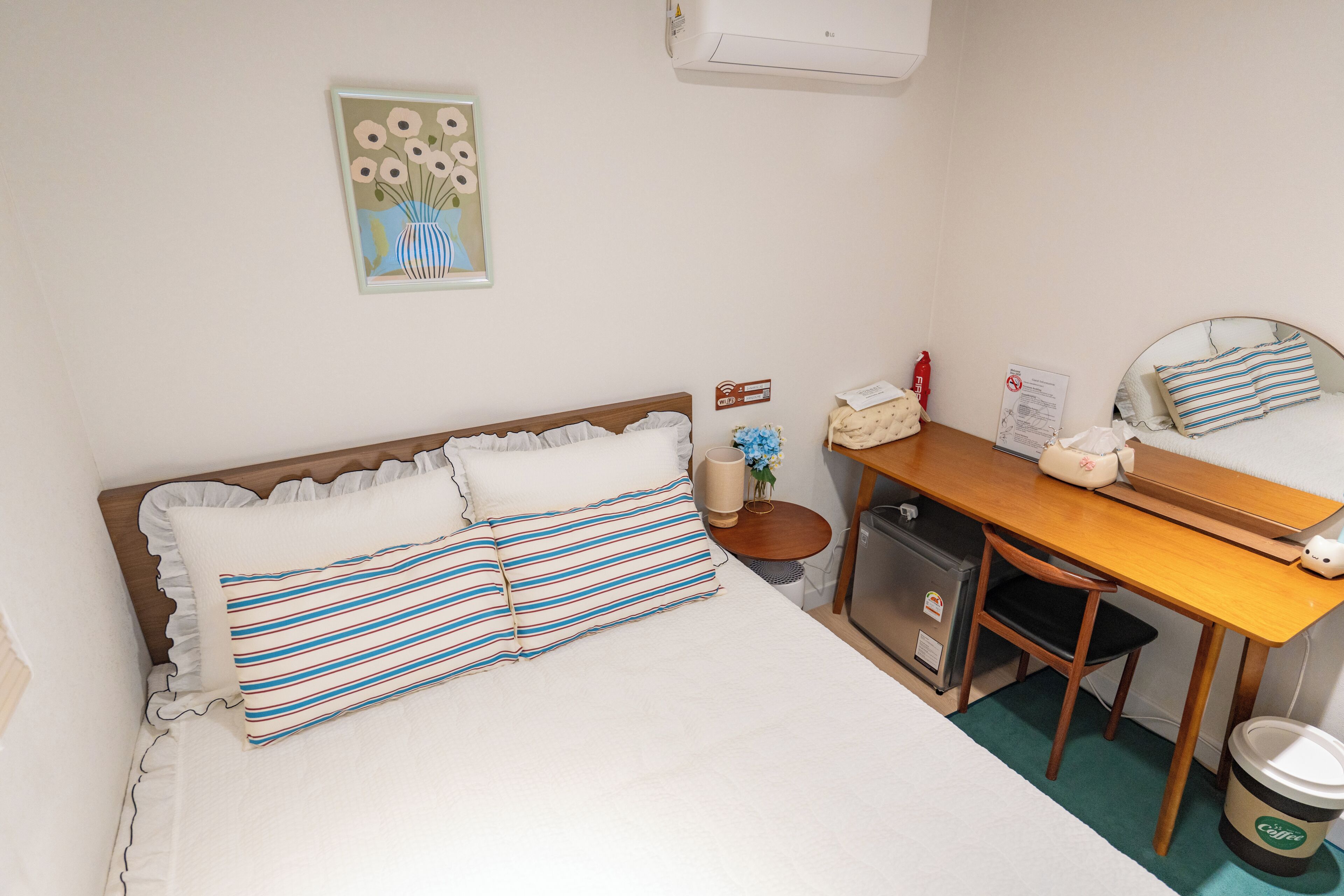 A neatly arranged hotel room with a bed, a desk, a chair, a small cabinet, and a trash bin.