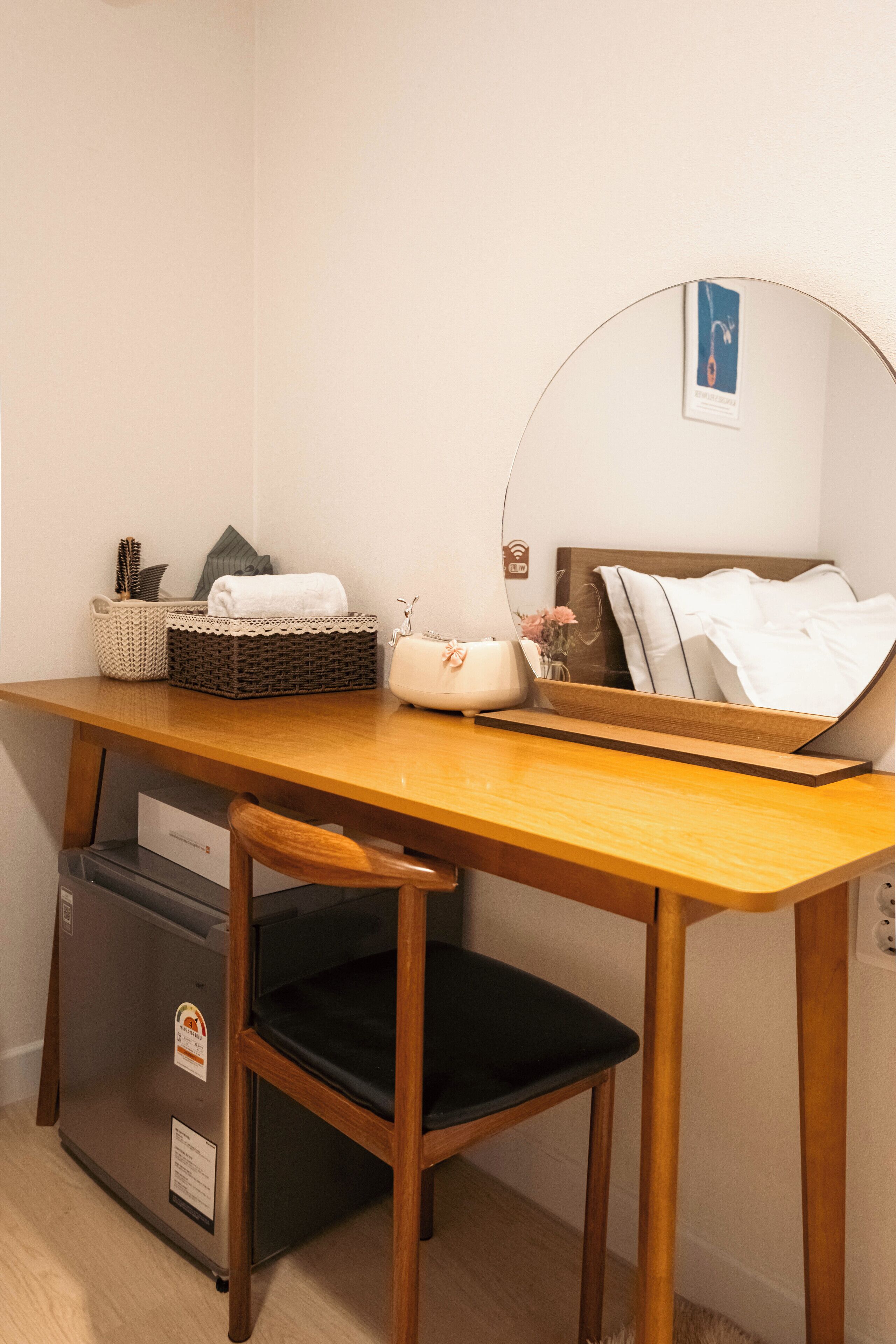 Une pièce de taille modeste, comprenant un bureau en bois, une chaise, un petit réfrigérateur, un miroir et une étagère fixée au mur, agrémentée d’un panier et d’un vase.