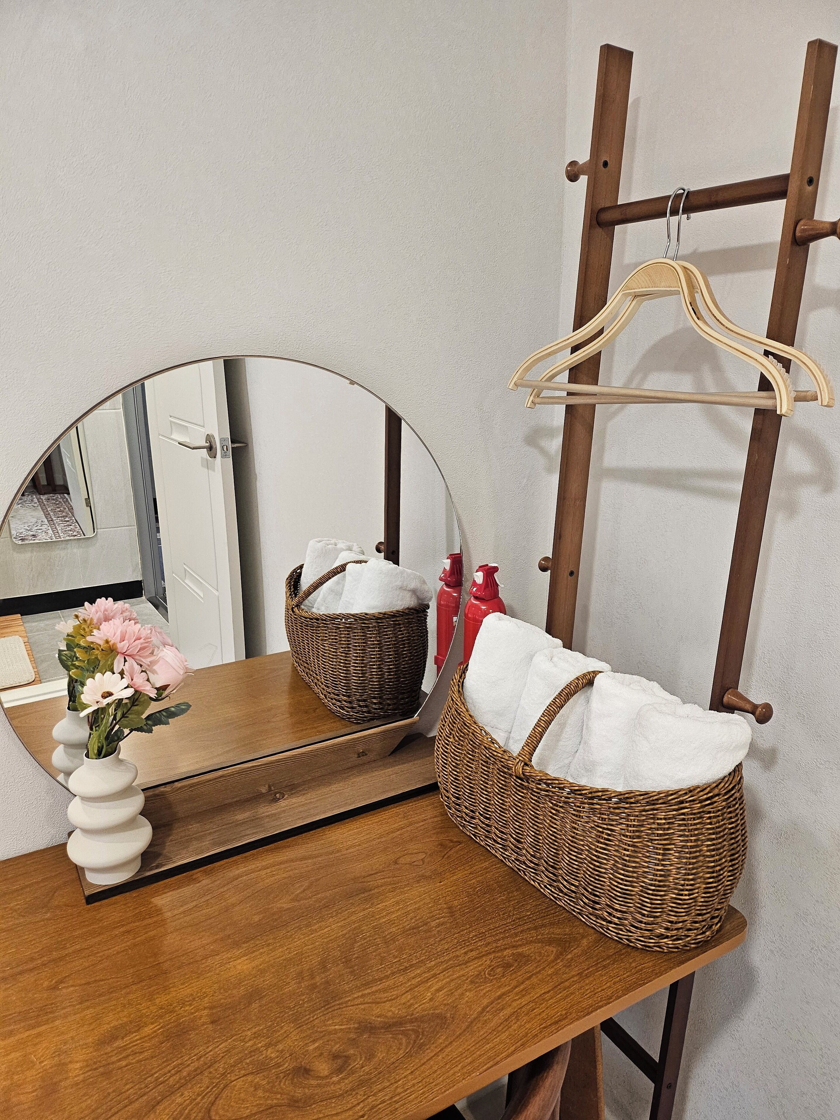 A wooden vanity with a round mirror, a wicker basket, and a vase with flowers.