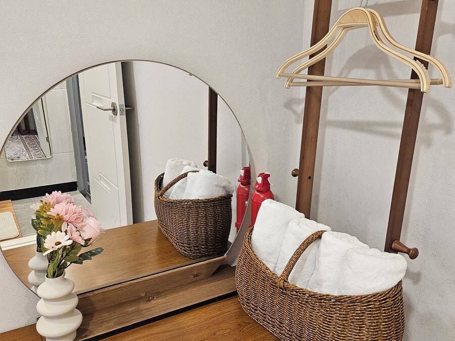 A wooden vanity with a round mirror, a wicker basket, and a vase with flowers.
