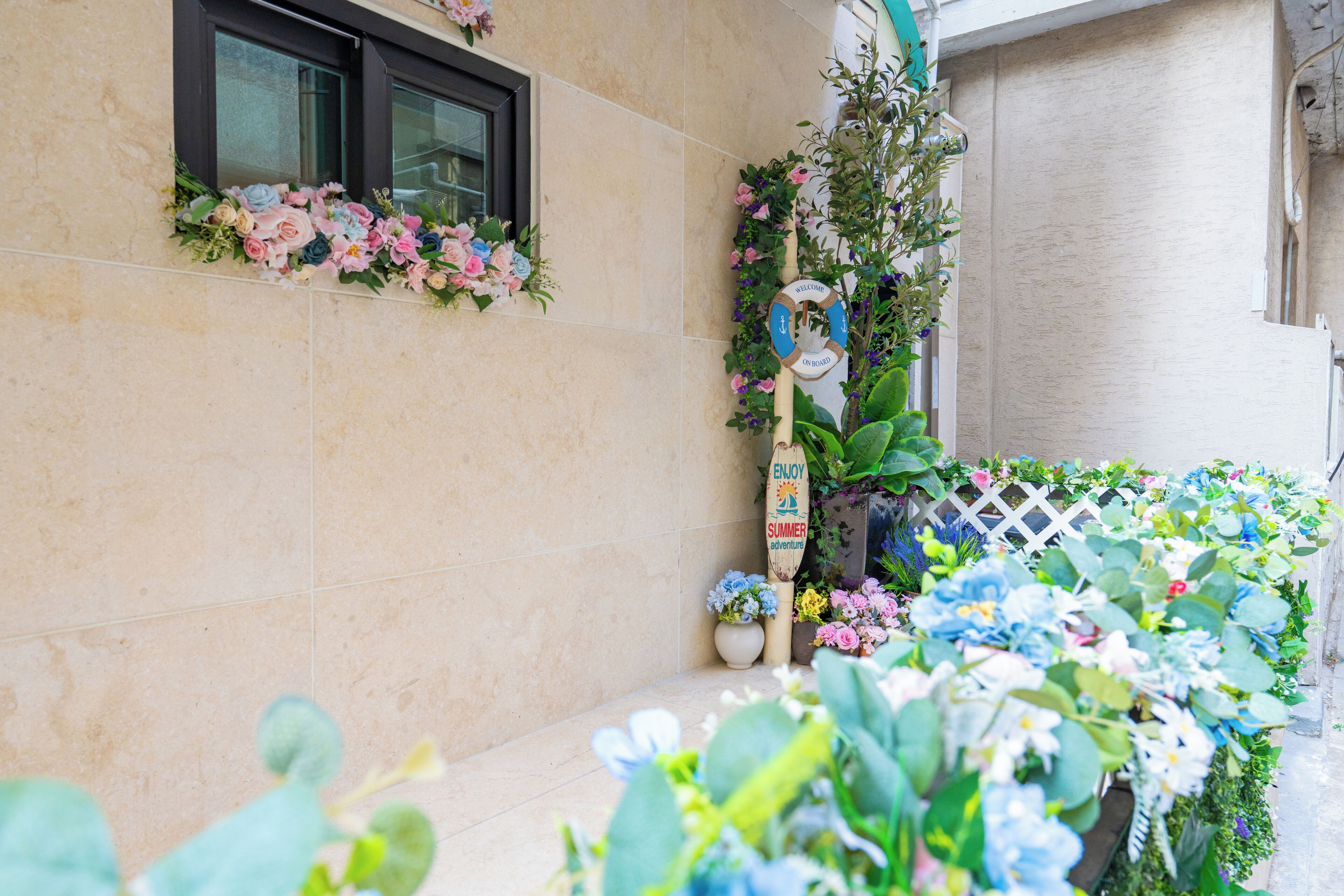 Eine Gebäudefassade mit einem Fenster, einem Willkommensschild und verschiedenen Kübelpflanzen und Blumen.
