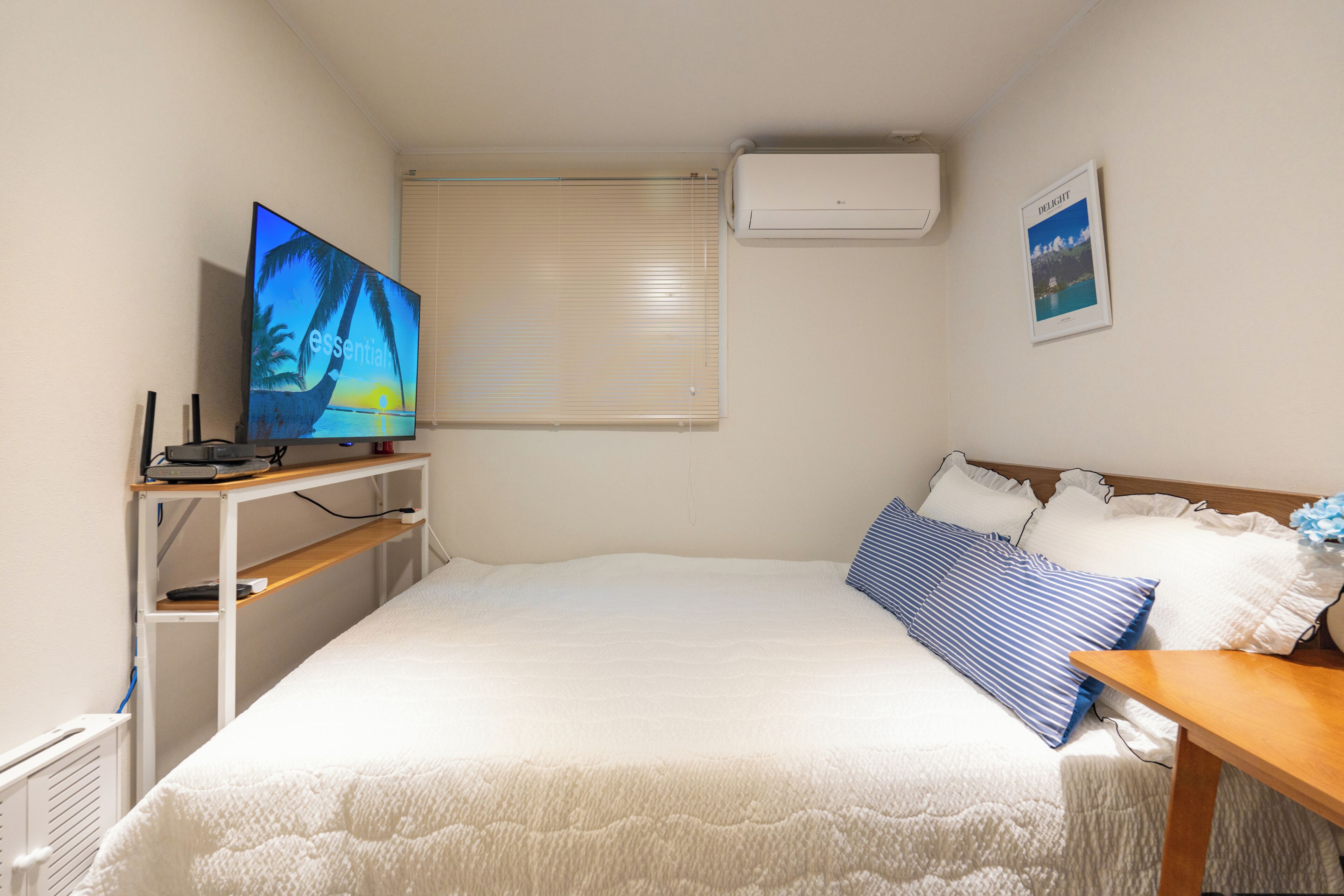 A bedroom with a bed, a TV, a window with blinds, an air conditioner, and a framed picture on the wall.