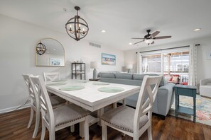 A dining area with a white table, chairs, and a chandelier. There is a gray sofa and a ceiling fan in the background. - DOG FRIENDLY & ALL NEW-SLEEPS 10-POOL CMMTY ON CANAL-21 Canal Landing, Rehob. (Rehoboth Beach)