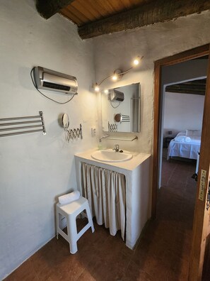 Ein Badezimmer mit einem weißen Waschbecken, einem Spiegel und einem Handtuchhalter. Ein kleiner Stuhl und ein Duschbereich sind im Hintergrund zu sehen.