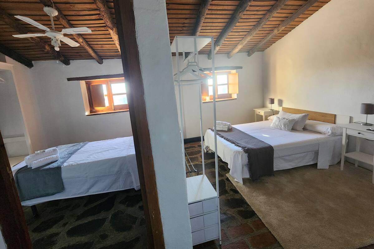 Una habitación con dos camas, techo de madera, suelo de piedra y una ventana con luz solar.