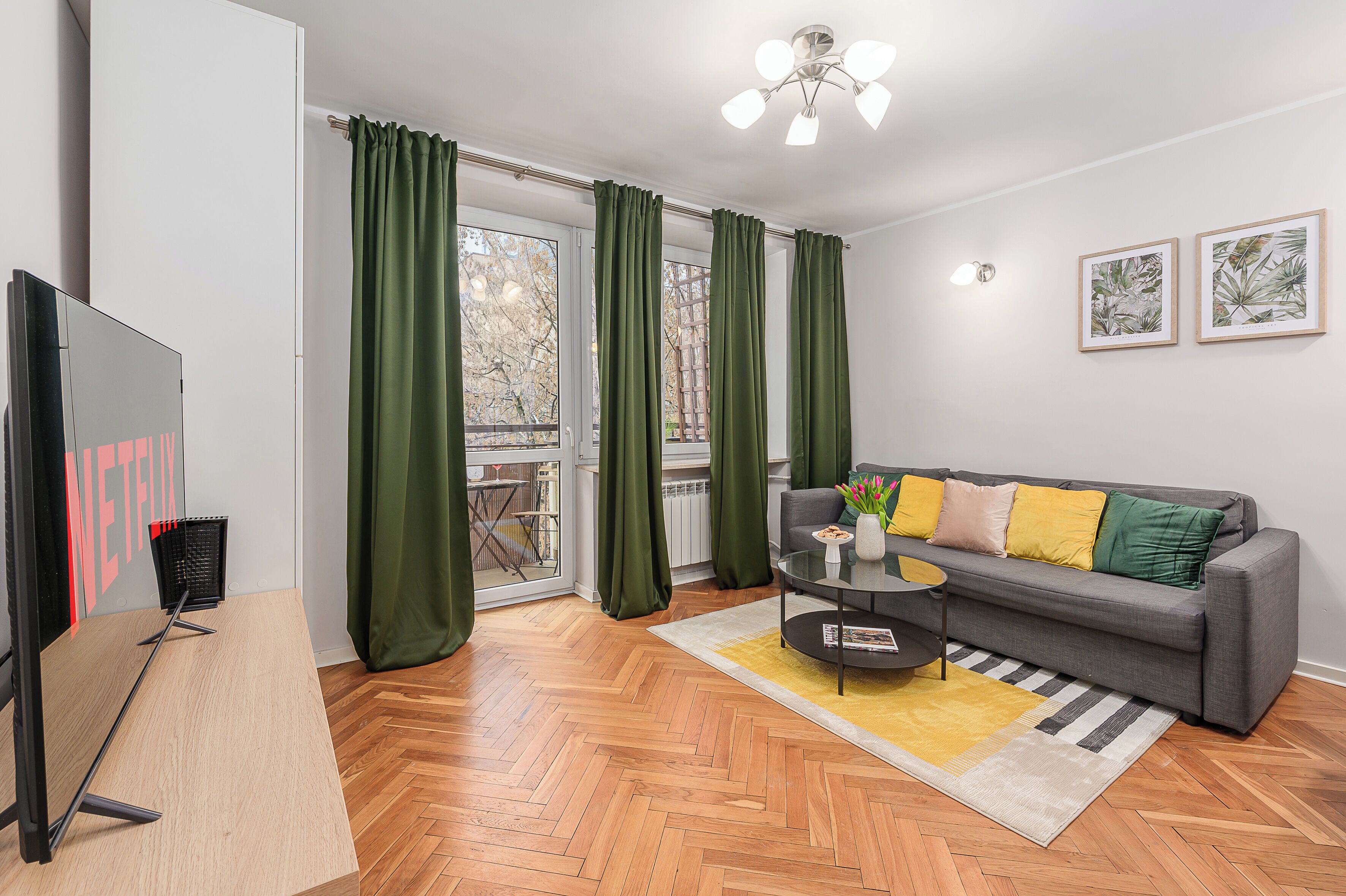 A modern living room with a grey sofa, green curtains, a wooden herringbone floor, and a flat-screen TV.