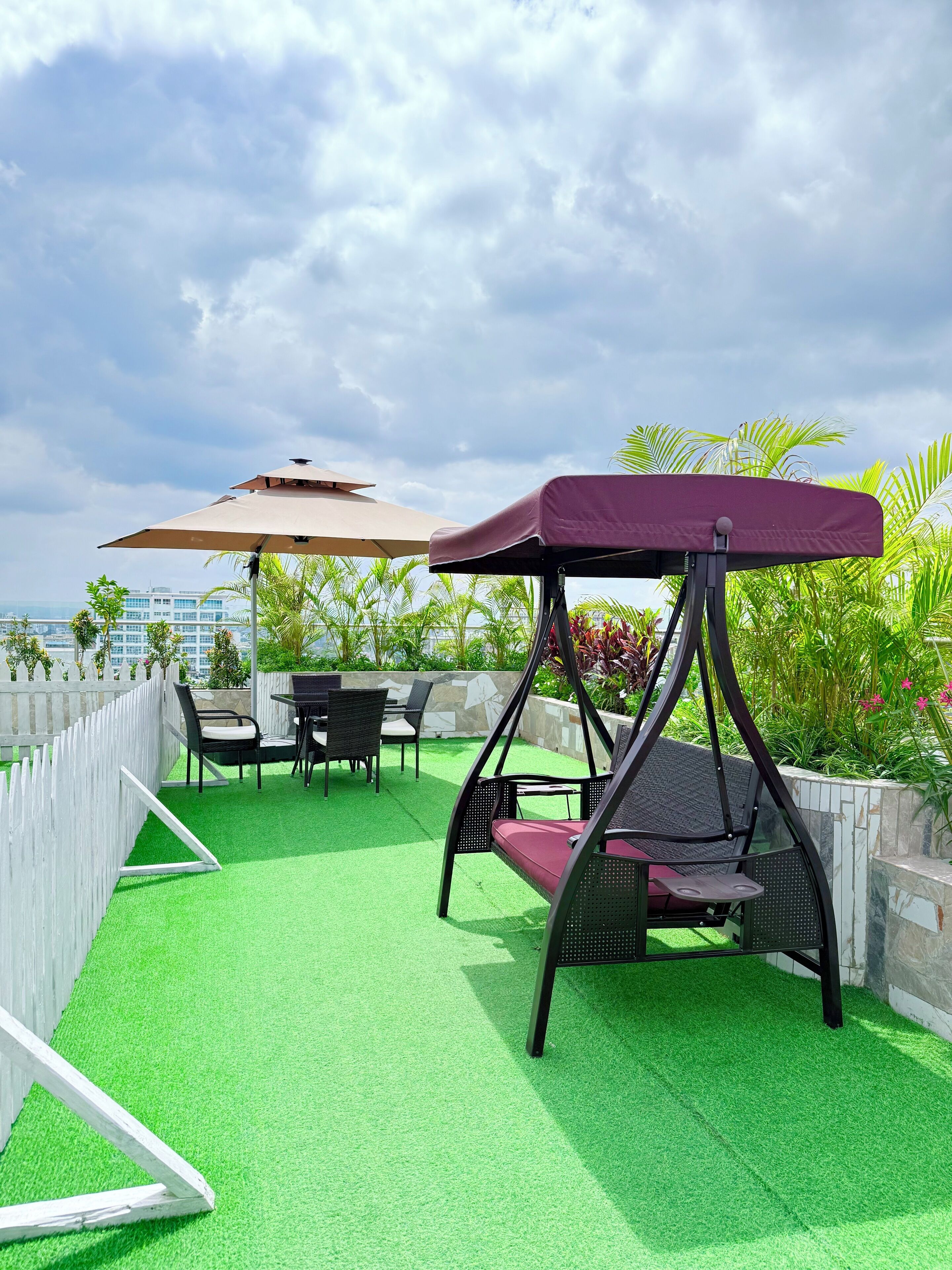 A patio area with green artificial turf, a swing chair, a table with chairs, and a white picket fence.