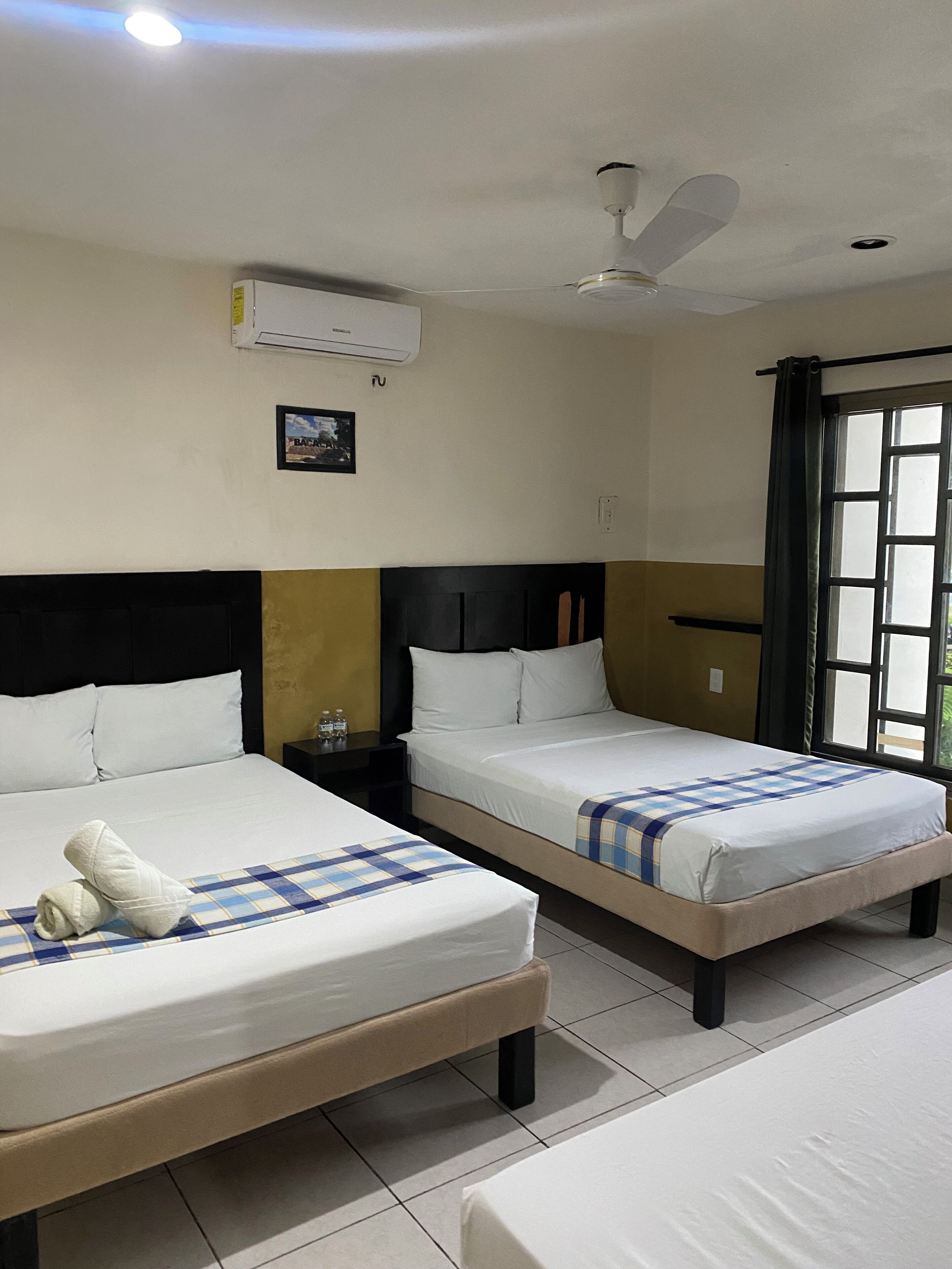 Quarto de hotel com duas camas, um ventilador de teto, ar condicionado, mesa de cabeceira e janela com cortinas.