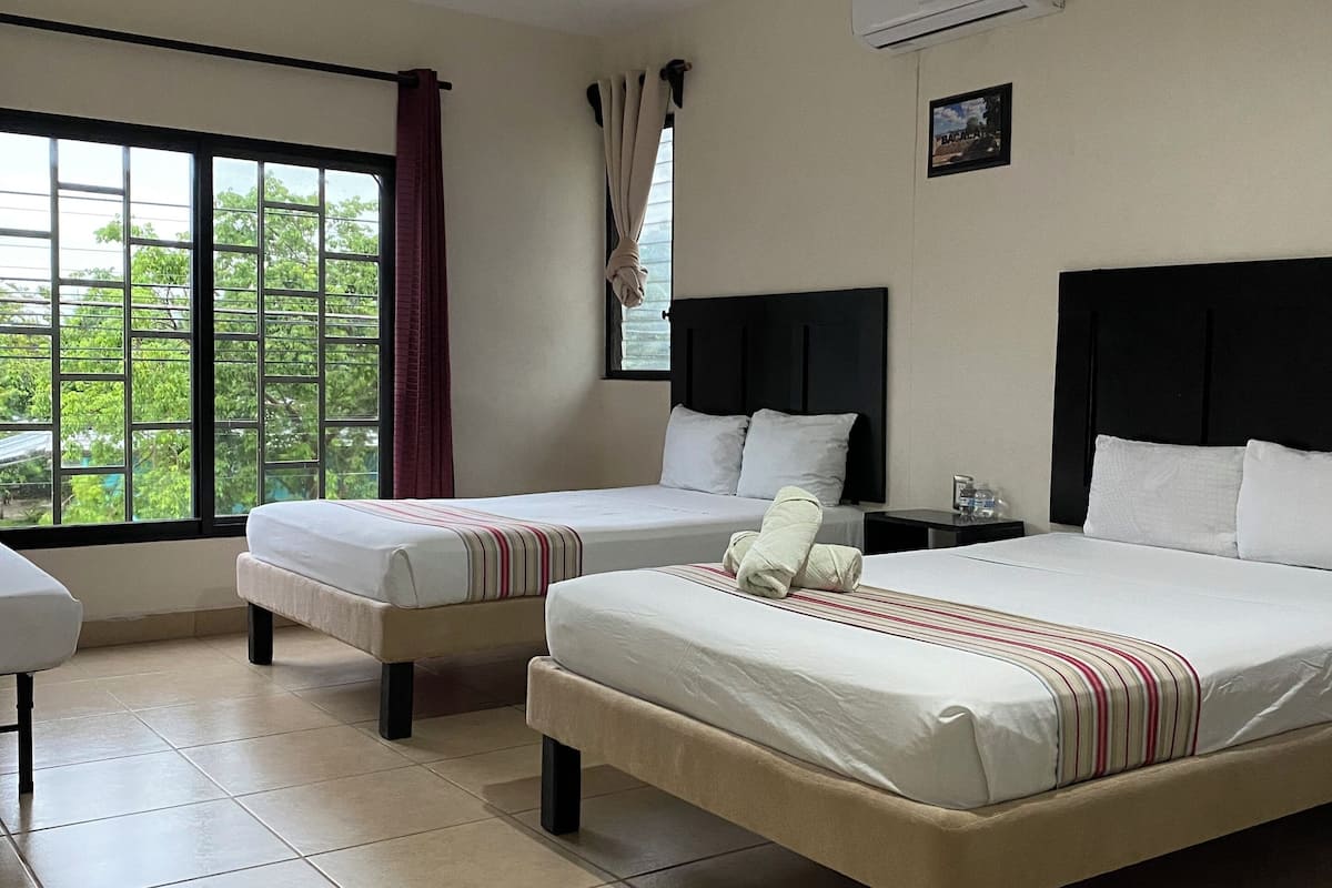 Ein Hotelzimmer mit zwei Betten, einem Deckenventilator, einem Fenster mit Vorhängen und Blick auf Grünfläche.