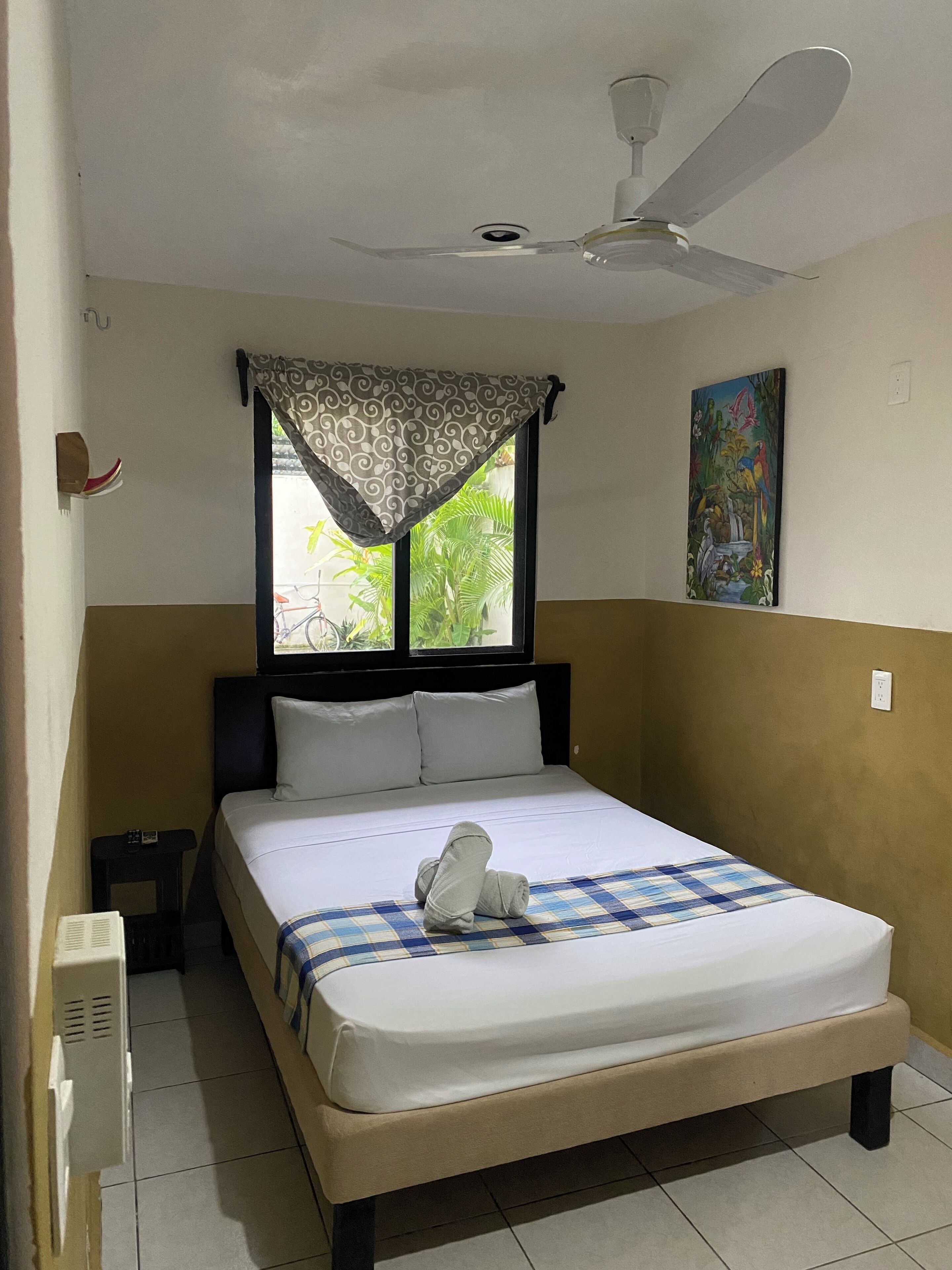 A neatly made bed with a checkered blanket, a ceiling fan, a window with a patterned curtain, and a painting on the wall.
