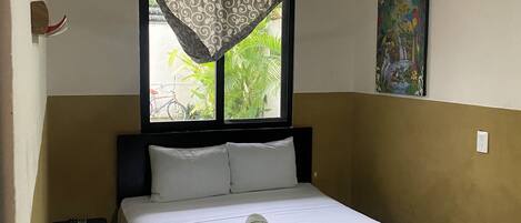 A neatly made bed with a checkered blanket, a ceiling fan, a window with a patterned curtain, and a painting on the wall.