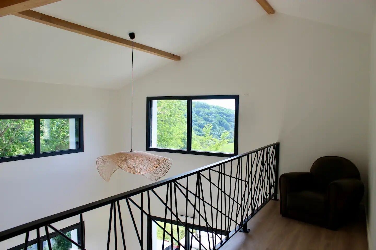 Une pièce spacieuse avec un plafond haut, un luminaire suspendu, une vue sur la verdure grâce à de grandes fenêtres et une rambarde noire.