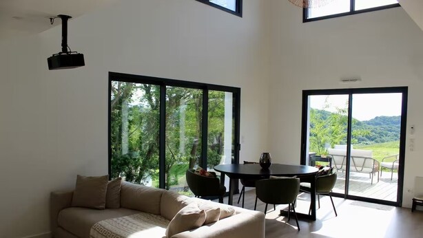 A spacious living room with a large window, a sofa, a dining table with chairs, and a view of a lush garden. - Pays Basque: Family home with large garden, close to mountains and ocean! (Mouguerre)