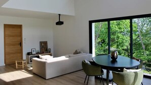 A modern living room with a white sofa, a dining table with chairs, and a large window with a view of trees. - Pays Basque: Family home with large garden, close to mountains and ocean! (Mouguerre)