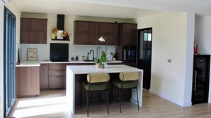 A modern kitchen with wooden cabinets, a central island with chairs, a refrigerator, and a dining area. - Pays Basque: Family home with large garden, close to mountains and ocean! (Mouguerre)