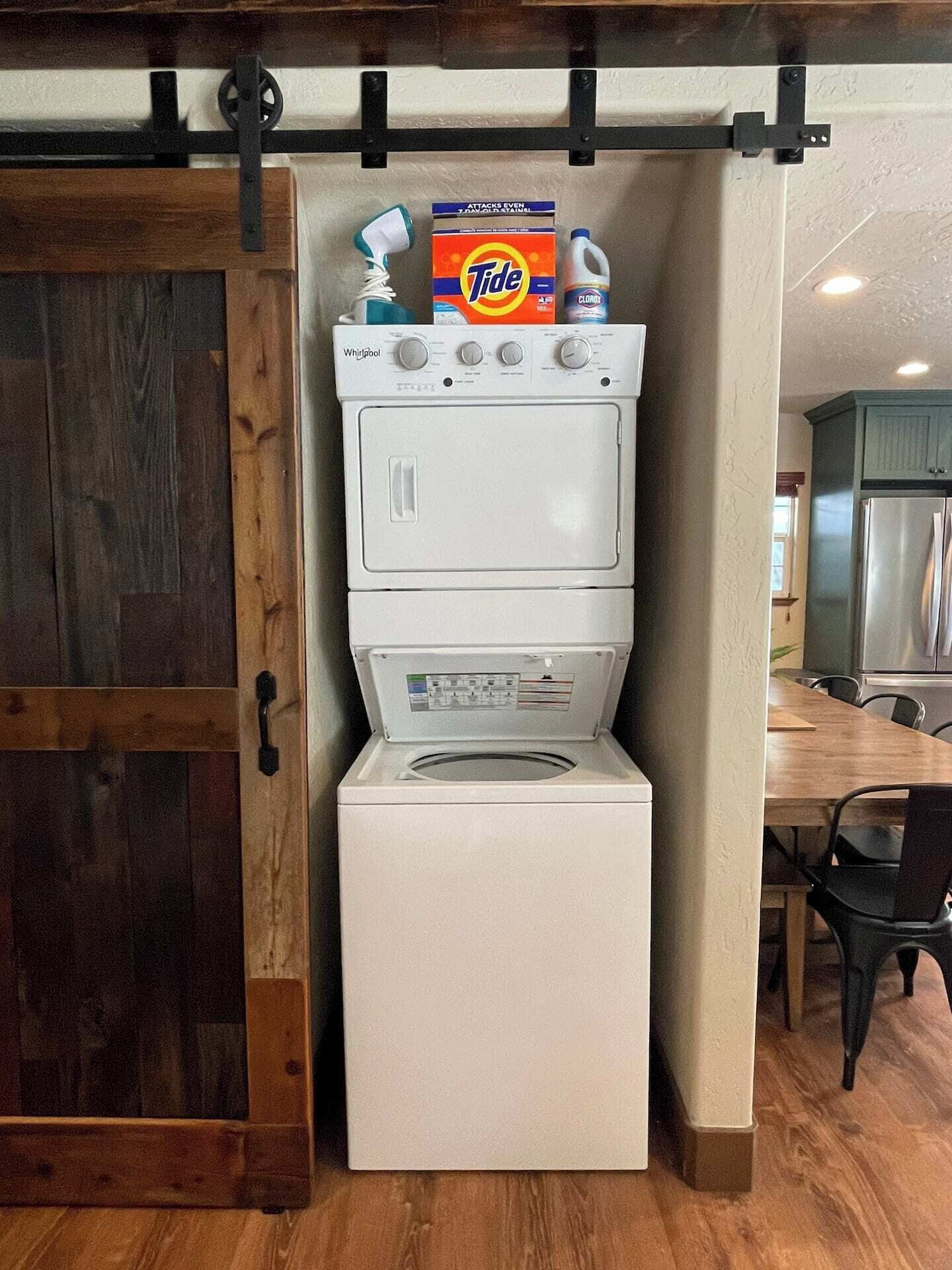 A compact laundry room with a built-in washer and dryer, a wooden door, and a dining area visible in the background.