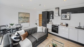 A modern apartment with a grey sofa, dining table, and chairs, a kitchen with white cabinets and appliances, and a painting on the wall. - York Suite (Chester)
