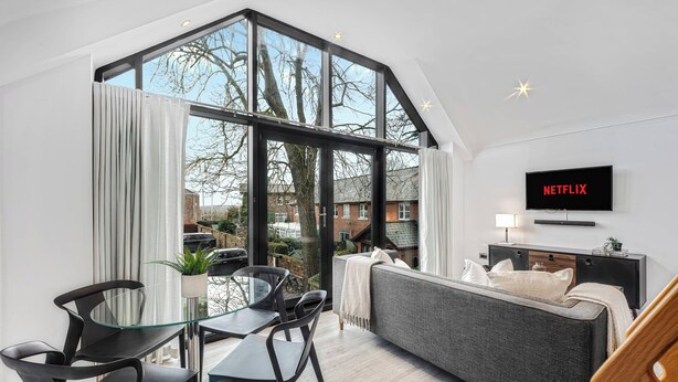 Ein modernes Wohnzimmer mit großem Fenster, einem grauen Sofa, einem an der Wand montierten Fernseher und einem Essbereich mit schwarzen Stühlen und einem Glastisch.