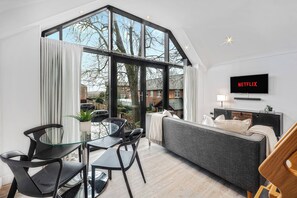 A modern living room with a large window, a grey sofa, a TV mounted on the wall, and a dining area with black chairs and a glass table. - Grosvenor Apartment (Chester)