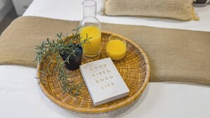 Ein Frühstücksbrett mit einem Buch mit dem Titel „Good Vibes, Good Life“ von Vick King, einem Glas Orangensaft, einem weiteren Glas Orangensaft und einer Topfpflanze auf einem Bett mit einem beigen Kopfkissen.