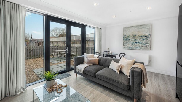 A modern living room with a grey sofa, a glass coffee table, and a large sliding glass door leading to a backyard. - Newland Suite (Chester)