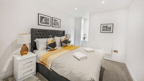 A modern bedroom with a tufted headboard, a bedside table with a lamp, and a bed with a white and yellow blanket. - Newland Suite (Chester)