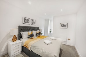 A modern bedroom with a tufted headboard, a bedside table with a lamp, and a bed with a white and yellow blanket. - Newland Suite (Chester)