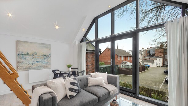 Ein modernes Wohnzimmer mit grauem Sofa, einem Glastisch und einem großen Fenster mit Blick auf eine Wohngegend.