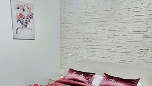 Un lit bien fait, recouvert d’une couette rouge, avec des oreillers brodés des mots « Famille » et « Maison », et des serviettes ornées de motifs floraux.