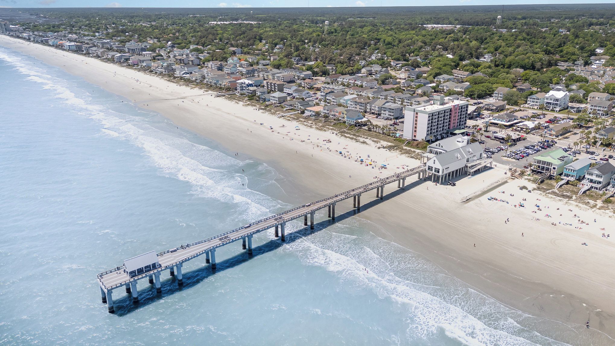 An aerial view of a coastal town with a long pier extending into the ocean, connecting the beach to the town.