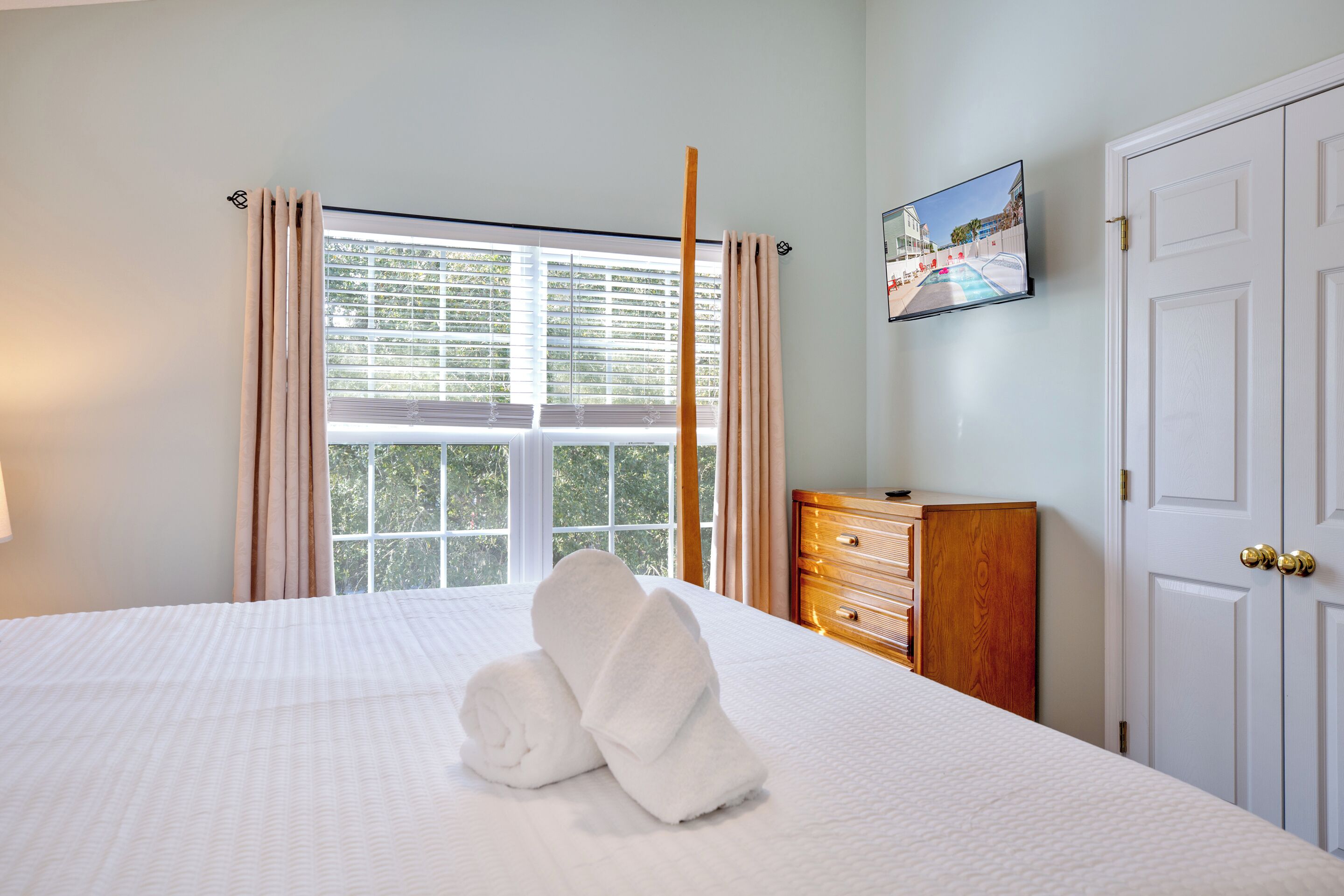 A neatly made bed with rolled towels, a wooden dresser, a television on the wall, and a white door.