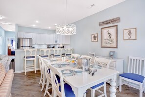 A modern kitchen with white cabinetry, a stainless steel refrigerator, and a dining area with white and blue chairs. - Palmetto Breeze Coastal Retreat w Private Pool (Surfside Beach)