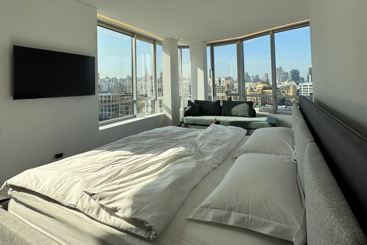 A modern bedroom with a large bed, a sofa, and a TV. It features large windows with a city view.