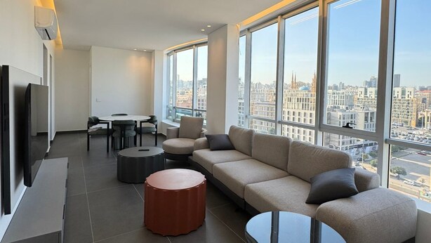 Un salón moderno con un sofá grande, una mesa de centro y un televisor. Hay ventanas amplias con vistas a la ciudad.
