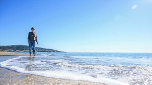 A person walking on a pebble beach with calm sea waves, a clear sky, and a distant shoreline. - VILLA des Rêves, Superbe Vue, Swimming Pool, Prox center (Sète)