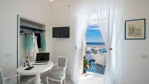 Une pièce avec un ordinateur portable sur une table, un téléviseur accroché au mur et une vue sur la mer depuis un balcon.