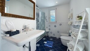 Une salle de bain avec un lavabo blanc, un miroir et des toilettes. Il y a une fenêtre et une étagère avec des serviettes.