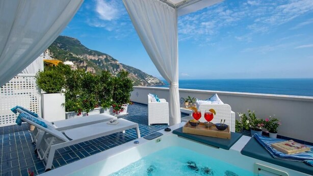 Un espace piscine aménagé sur le toit, offrant une vue sur la mer et les montagnes, avec des chaises longues blanches et des plantes en pot.