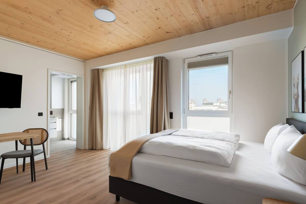Habitació d'hotel moderna amb sostre de fusta, un llit gran amb roba de llit blanca, un escriptori amb una cadira i vista a la ciutat per la finestra.