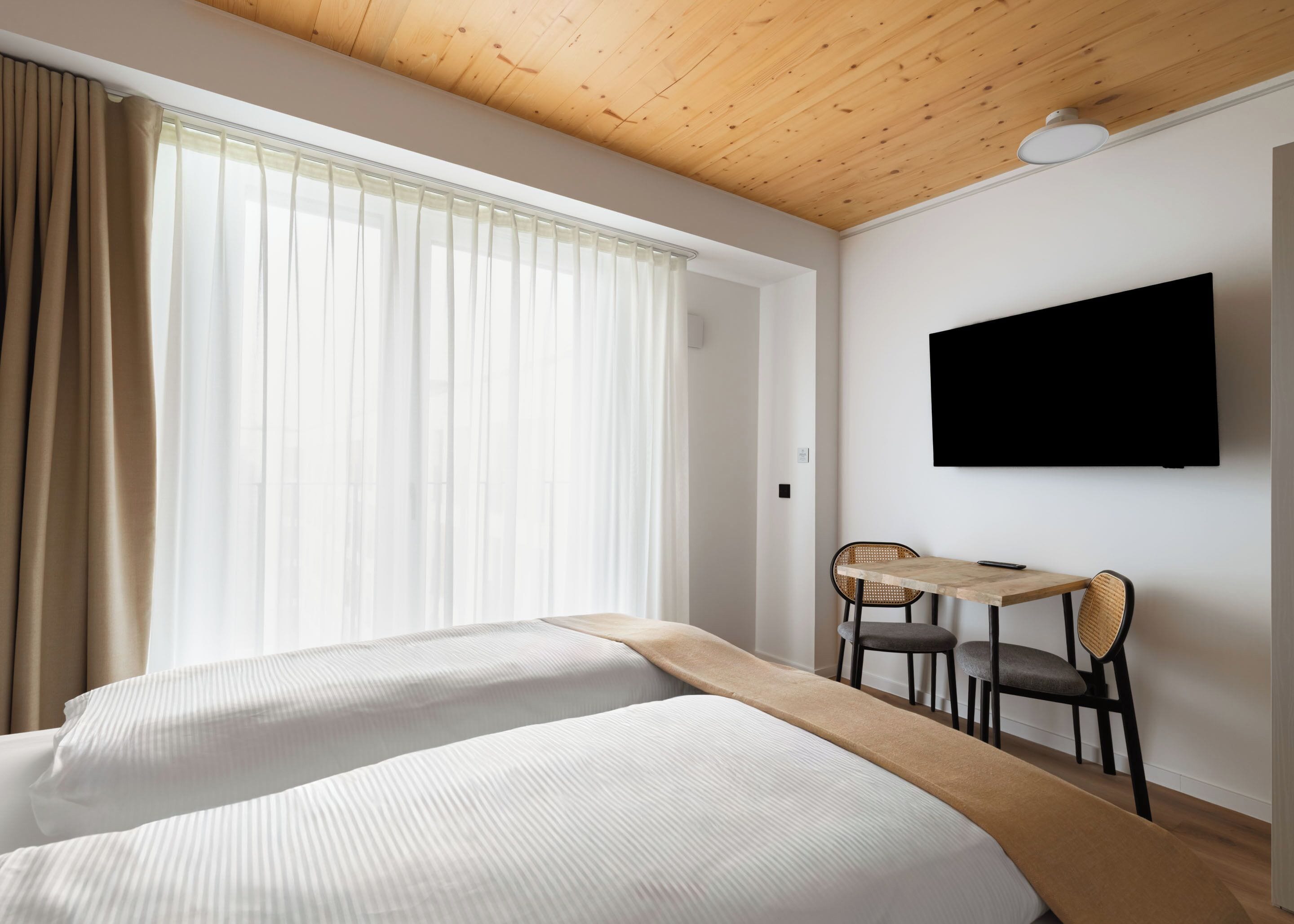 Hotelkamer met twee bedden, een houten plafond, een aan de muur bevestigde televisie en een raam met gordijnen.