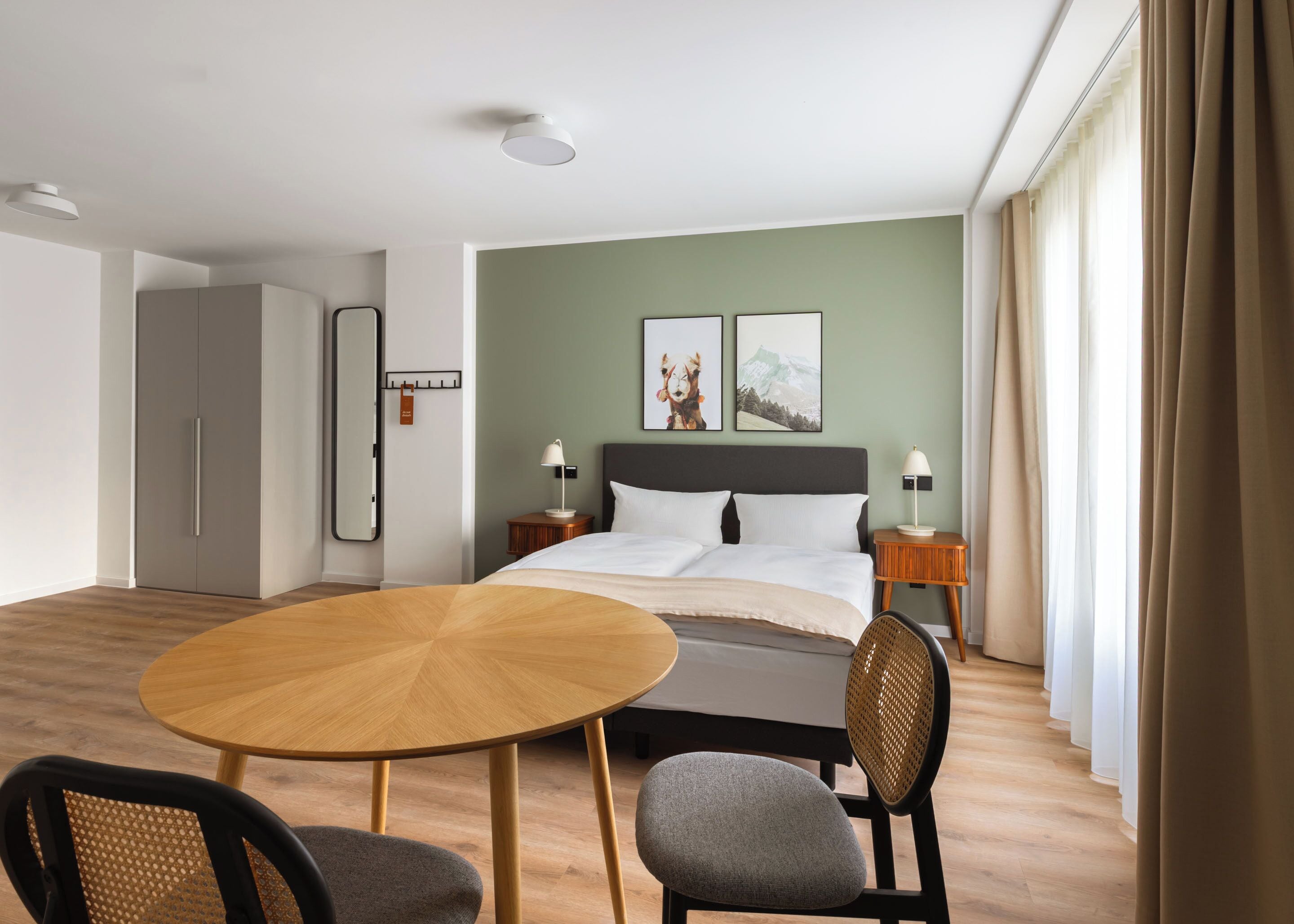 A hotel room with a bed, bedside tables, a round wooden table, two chairs, a mirror, and artwork on the wall.