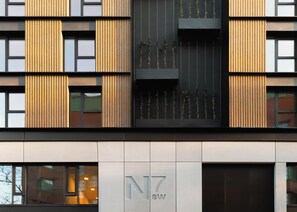 A modern building facade with vertical wooden slats, a black vertical panel with plants, and a sign with "NZ" and "ew" indicating the name.