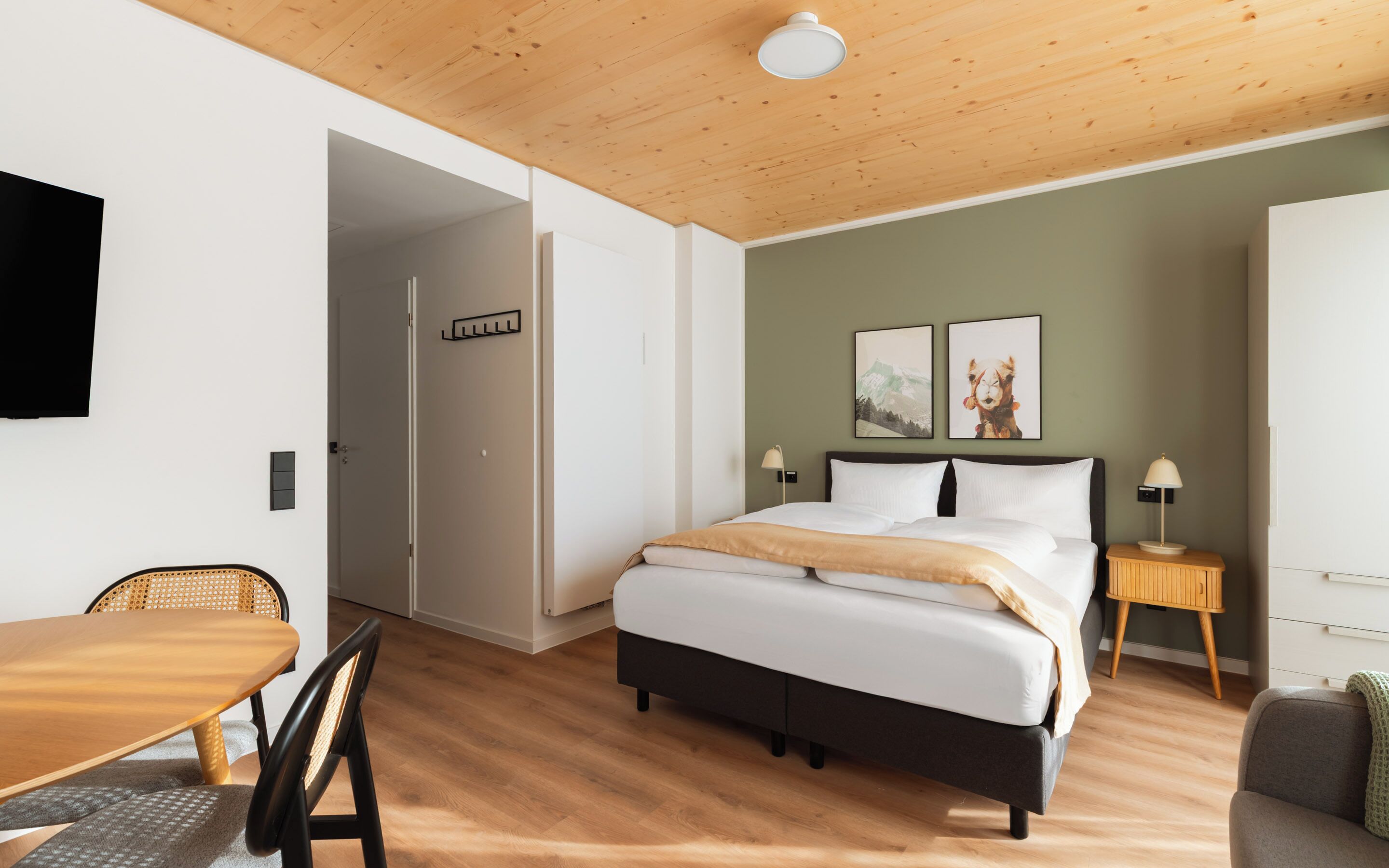 A modern bedroom with a wooden ceiling, a bed with white bedding, a bedside table with a lamp, and two framed pictures on the wall.