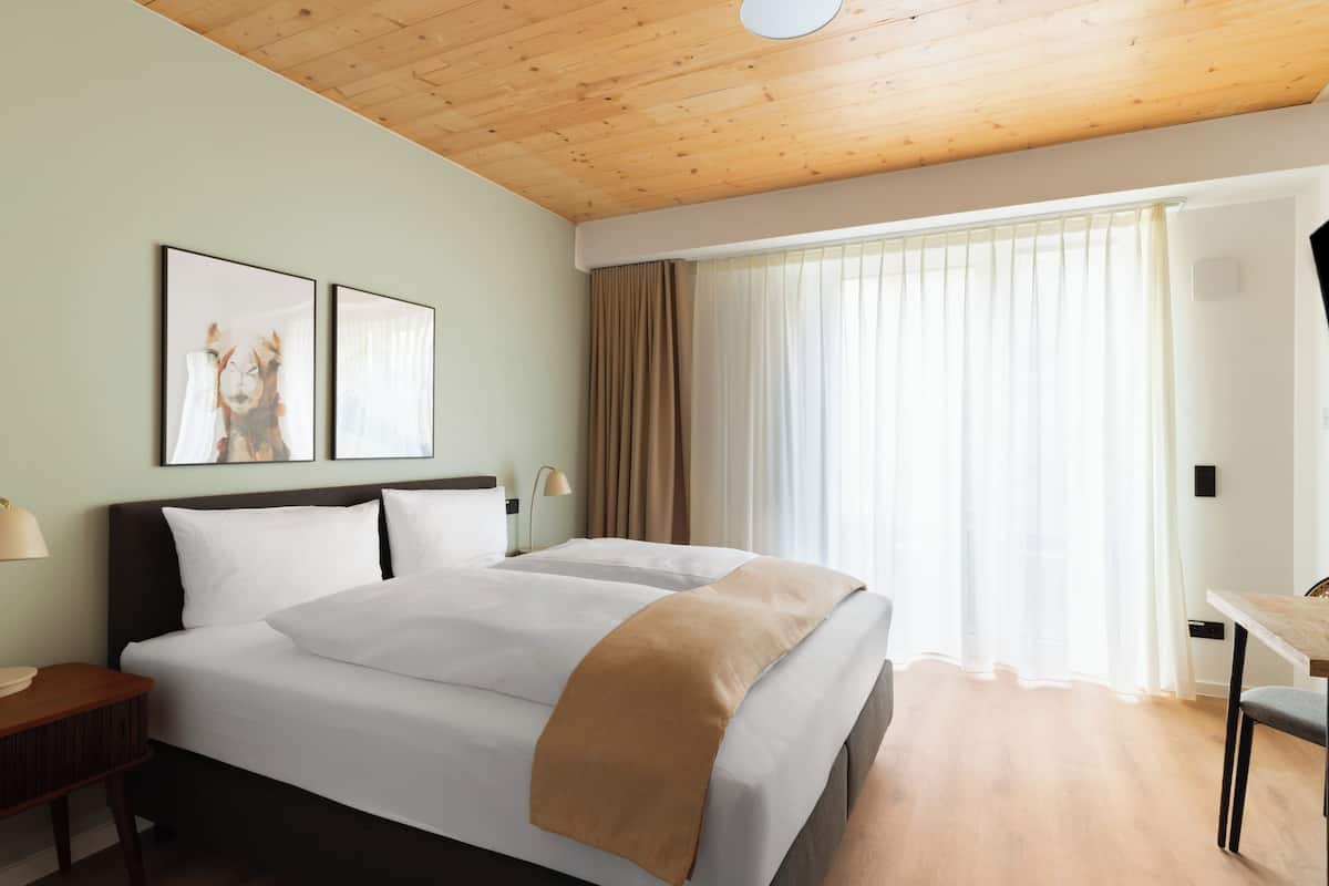 Habitació d'hotel amb sostre de fusta, un llit gran amb roba de llit blanca, una taula de nit amb una làmpada i una finestra amb cortines.