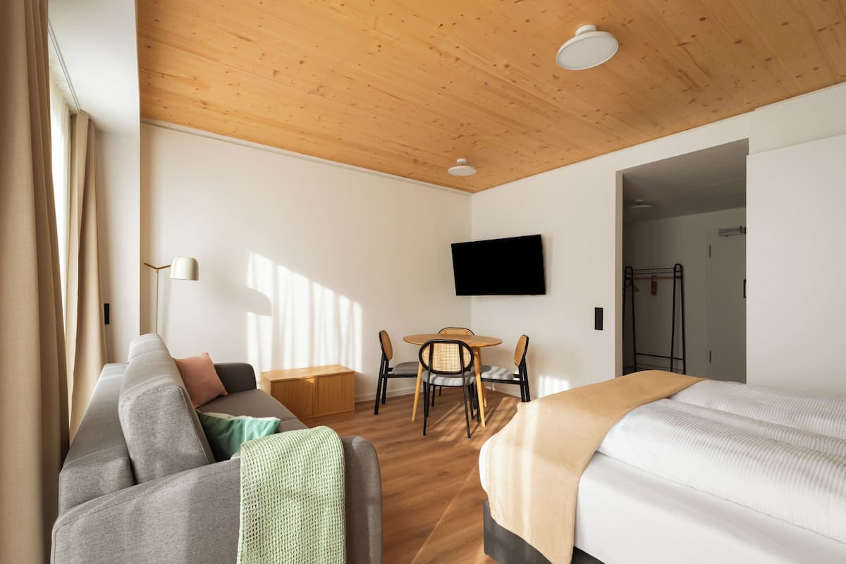 Habitació d'hotel moderna amb sostre de fusta, televisió de pantalla plana penjada a la paret, un sofà, un llit, taula de menjador amb cadires i un petit armari de fusta.