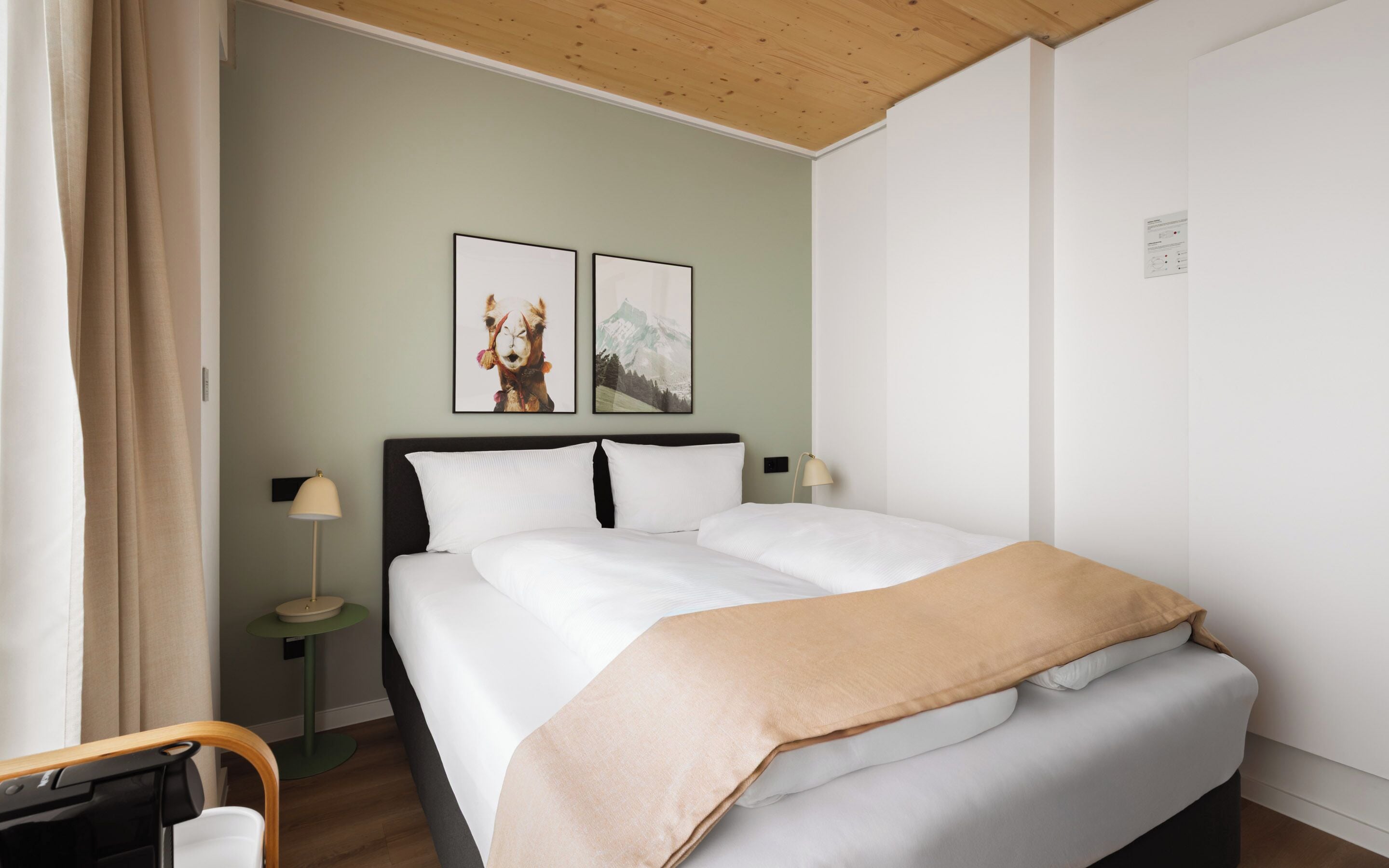Een net opgemaakt bed met witte lakens en een beige plaid, een klein nachtkastje met een lamp, en twee ingelijste foto's aan de muur.