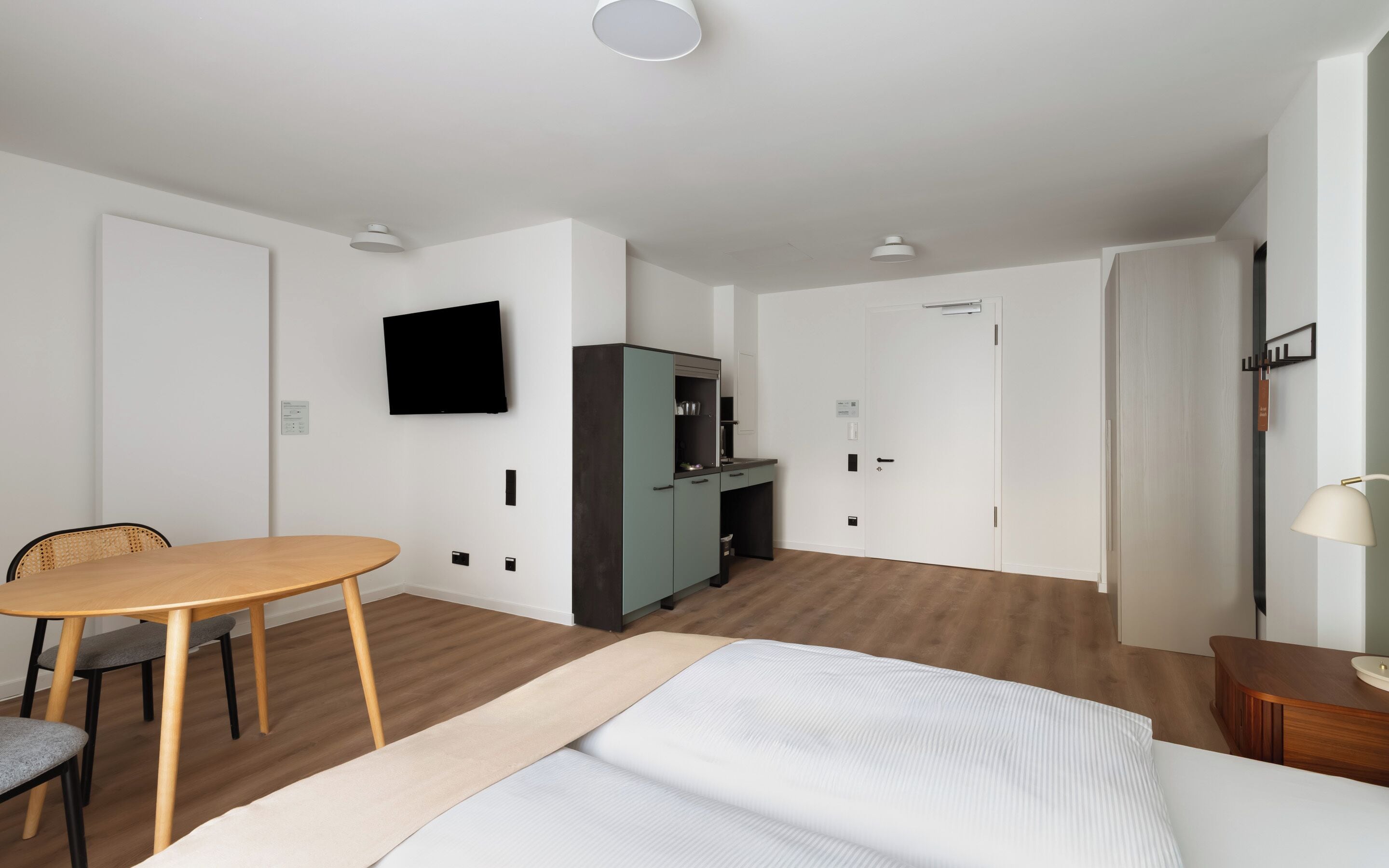 A modern hotel room with a bed, a desk, a chair, a television, and a wardrobe.
