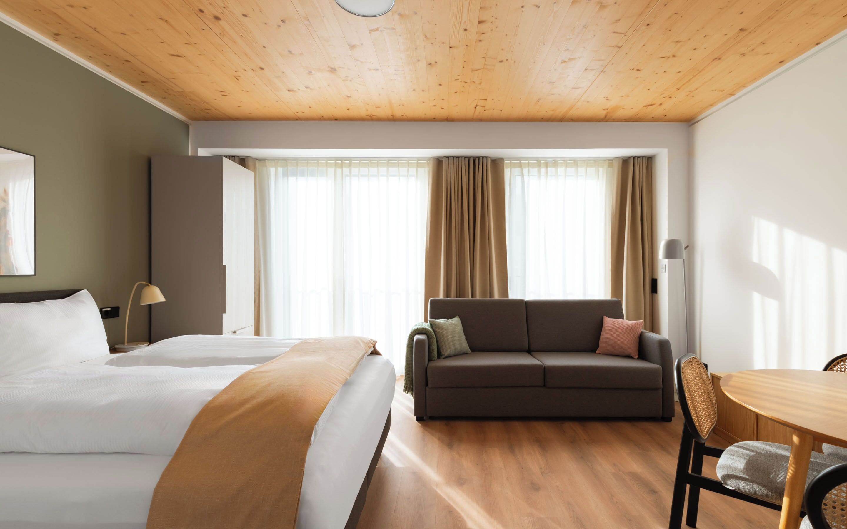 Ein Hotelzimmer mit Holzboden, einem Bett mit weißen Leinentüchern, einem braunen Sofa, einem Esstisch und einem Stuhl.