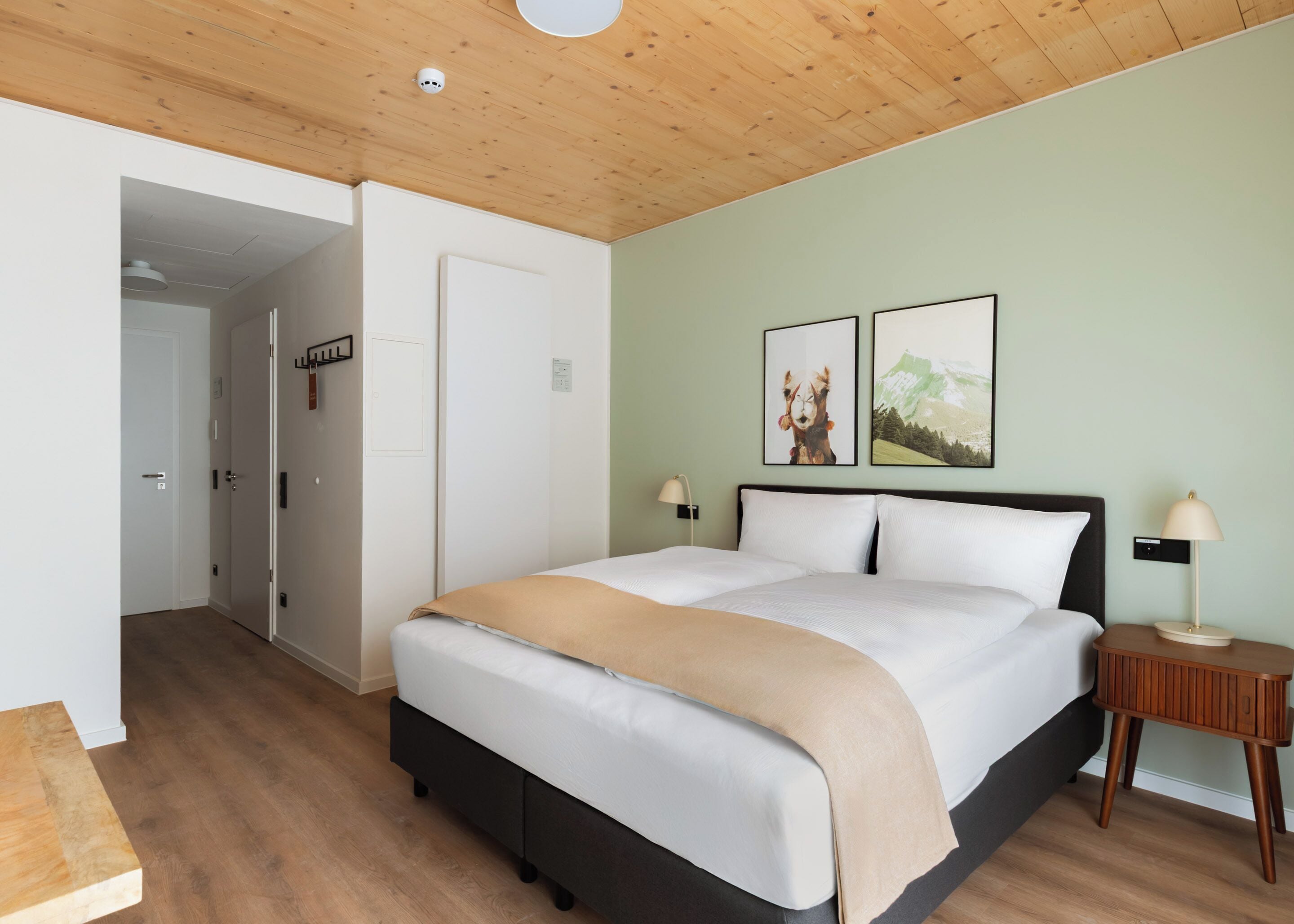 Un dormitorio con cama, mesitas de noche, una puerta y dos cuadros en la pared.